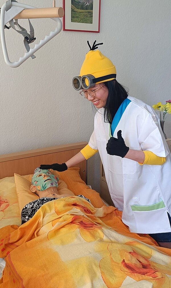 Operation Krankmacher: Die Mission der Minions Eine junge Frau im Minions-Kostüm mit Pflegekleidung und schwarzen Handschuhen steht an einem Pflegebett, in dem eine Pflegeübungspuppe mit Gesichtsmaske liegt. Die eine Hand hat die Schülerin auf den Kopf der Pflegepuppe gelegt und mit der anderen zeigt sie den Daumen hoch.