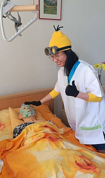 Eine junge Frau im Minions-Kostüm mit Pflegekleidung und schwarzen Handschuhen steht an einem Pflegebett, in dem eine Pflegeübungspuppe mit Gesichtsmaske liegt. Die eine Hand hat die Schülerin auf den Kopf der Pflegepuppe gelegt und mit der anderen zeigt sie den Daumen hoch.