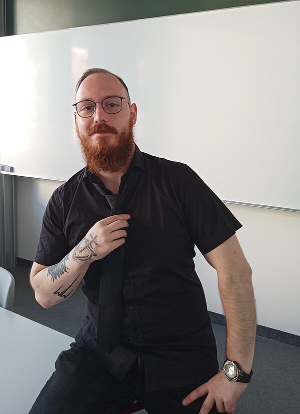 Alumni im Gespräch: Interview mit Florian Richter Absolvent Florian Richter sitzt auf einem Tisch im Unterrichtsraum, er trägt eine dunkle Hose, ein dunkles und kurzärmliges Hemd, eine Brille, eine Armbanduhr sowie einen Bart und lächelt in die Kamera.