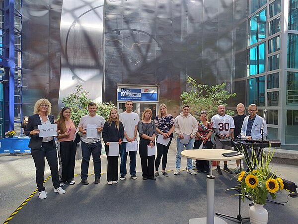 Neue Klassen in über einem halben Dutzend Bildungsgänge gestartet Eine Gruppe von zwölf Personen, darunter eine Frau mit einem Schild „Phy 25 TZ“, steht auf der Bühne mit Zertifikaten und kleinen Geschenken.