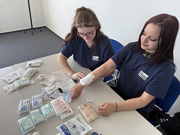 Tag der offenen Tür an der Euro Akademie Bitterfeld-Wolfen Zwei weibliche Auszubildende in marineblauen Euro Akademie-T-Shirts sitzen an einem Tisch, auf dem zahlreiche Verbände, Pflaster, Kompressen liegen und demonstrieren das Anlegen eines Verbandes.