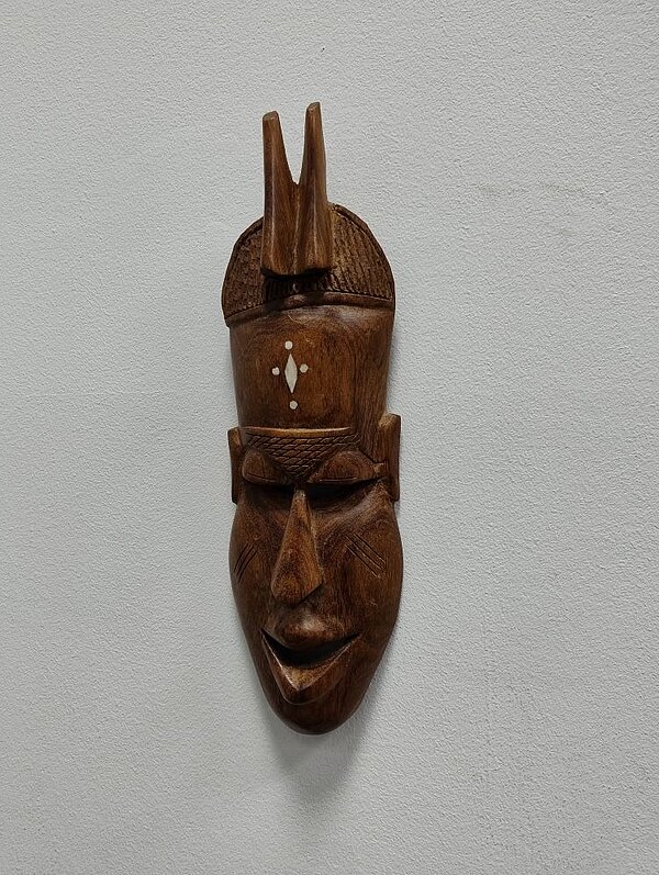 Eine dunkelbraune Holzmaske, auf der abstrakt ein Gesicht angedeutet ist, hängt an einer weißen Wand. Auf die Stirn ist ein weißes geometrisches Muster gezeichnet.