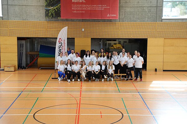 Auszubildende der Euro Akademie Pößneck unterstützen erneut den „Pokal des Bürgermeisters“ Die Schülerinnen und Schüler haben sich für ein Gruppenfoto vor die offenen Materialräume positioniert. Die vordere Reihe sitzt auf einer Holzbank, während die hintere Reihe steht. Eine Beachflag steht am Rand der Gruppe.
