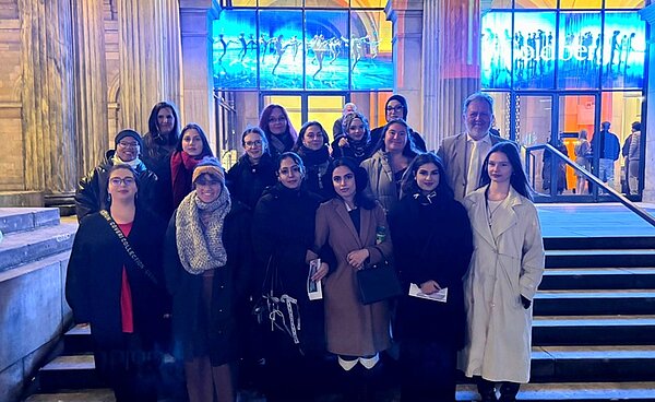 Die Schülerinnen und Schülern der Euro Akademie Hannover stehen vor dem beleuchteten Eingangsbereich auf einer Treppe des Opernhauses Hannover und posieren für ein Gruppenfoto. Sie tragen unterschiedliche Winterbekleidung, darunter Mäntel, Schals und Jacken in verschiedenen Farben wie Beige, Schwarz und Dunkelblau. Im Hintergrund sind große Glasfenster mit blauer Beleuchtung und der Schriftzug "Goldberg" sichtbar, der auf das Ballettstück hinweist, das sie besucht haben.