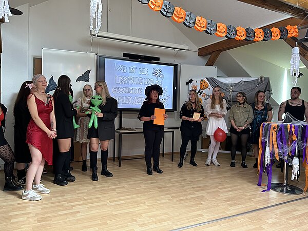 Kostümierte Gruppe steht auf einer Bühne im dekorierten Klassenraum; an der Leinwand erscheint die Frage „Was ist der Ursprung und die Geschichte von Halloween?“