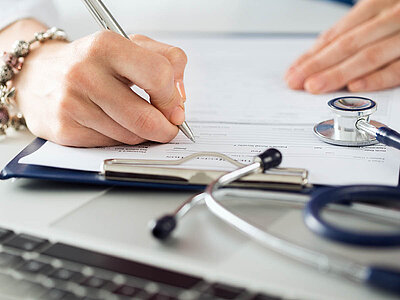 Das Bild zeigt jemanden, der ein medizinisches Formular ausfüllt, während ein Stethoskop und ein Laptop auf dem Tisch liegen. Es veranschaulicht den Zusammenhang zwischen Gesundheitsversorgung und Dokumentation.