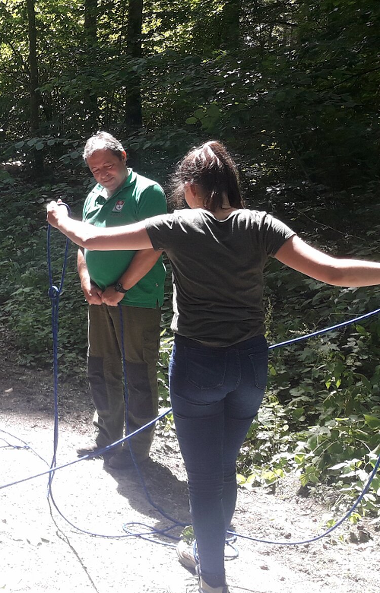 Waldtag des Berufskollegs Von hinten ist eine Schülerin zu erkennen, die mit beiden Händen ein blaues Seil festhält. Dabei schaut ihr der Förster prüfend zu. Die Szene spielt sich auf einem Waldweg ab.