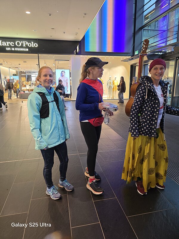 Drei Frauen in Sportkleidung stehen in einem Einkaufszentrum vor dem Marc O’Polo Store. Eine trägt eine Gitarre, die anderen zwei tragen Sportjacken und Laufkleidung.