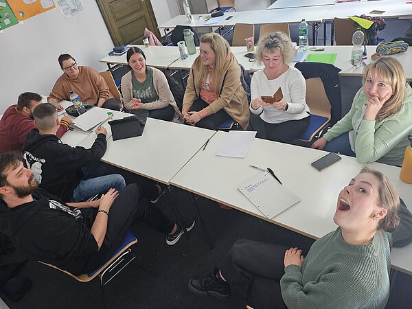 Podcast-Projekt: KAFFEE TRINKEN & SPIELEN Schülerinnen und Schüler der Euro Akademie Halle sitzen an einem langen Tisch und planen ihr Projekt, dabei schauen sie alle in die Kamera.