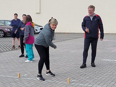 Auf einem Parkplatz spielen ein Mann und eine Frau auf der freien Fläche Wikingerschach. Auf dem Boden stehen kleine Hölzer, die Frau macht gerade einen Wurf. Im Hintergrund sind weitere Personen zu sehen.