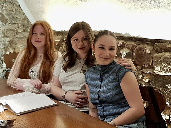 Alumnitreffen 2025 Drei weibliche ehemalige Auszubildende der Euro Akademie Aschaffenburg sitzen nebeneinander an einem Tisch im Restaurant und posieren lächelnd für ein Foto. Vor ihnen liegt eine Speisekarte.