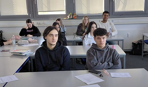 Mehrere Schüler*innen sitzen an ihren Tischen und verfolgen aufmerksam einen Vortrag im Klassenraum.