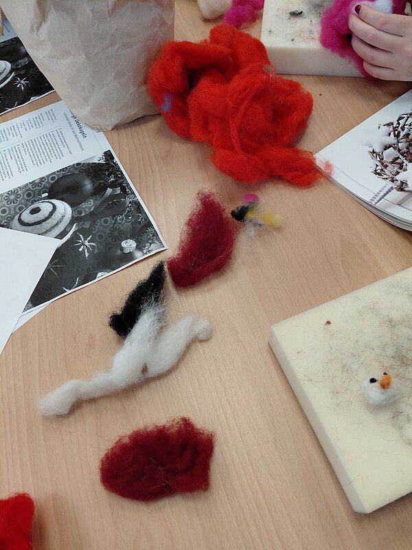 Bunte Wollfasern und kleine Filzteile liegen auf einem Tisch, daneben Arbeitsunterlagen – Materialien für eine Filzwerkstatt.