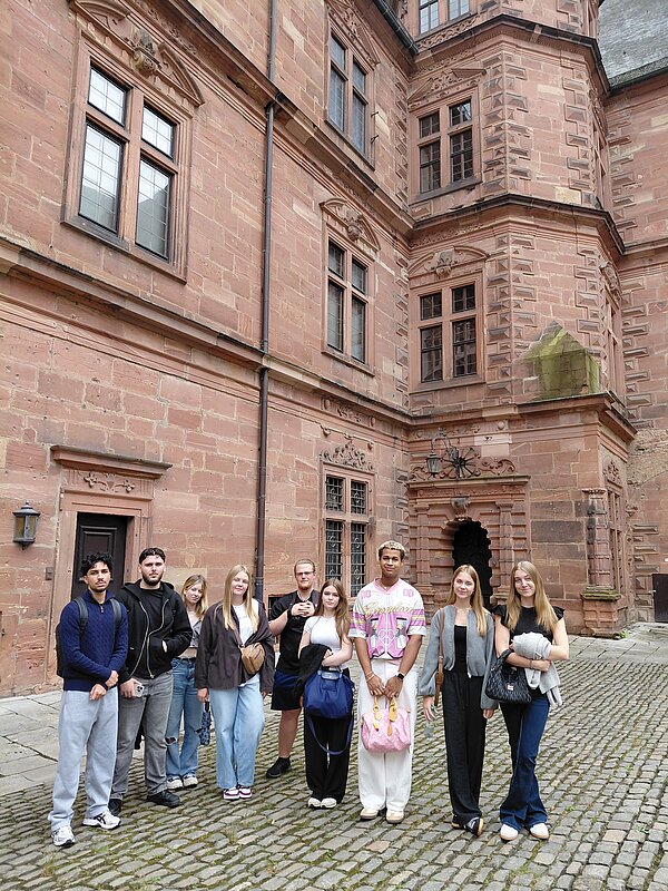 Eintauchen in die Geschichte Aschaffenburgs Eine Gruppe junger Menschen steht im Innenhof eines Schlosses aus rotem Sandstein. Die historische Fassade mit dekorativen Fenstern bildet den Hintergrund.