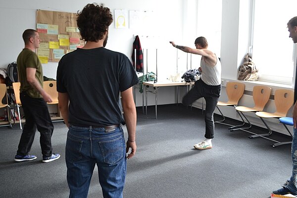 Vier Besucher des Tages der offenen Tür an der Euro Akademie Würzburg stehen in einem Klassenraum, in dem die Tische und Stühle zur Seite geschoben wurden und machen sportliche Übungen. 