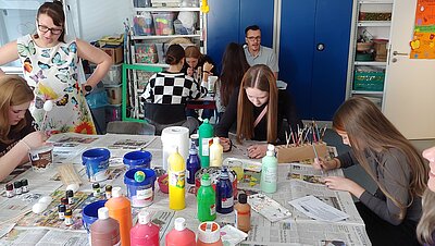 Kreative Bildung: Mehrere junge Frauen sitzen an zwei Tischen und bemalen mit Pinseln unterschiedliche Gegenstände. Auf den Tischen, die mit Zeitungspapier ausgelegt sind, stehen verschiedene Flaschen mit bunten Farben darin. Ein Mann hat sich auch an den hinteren Tisch verirrt.