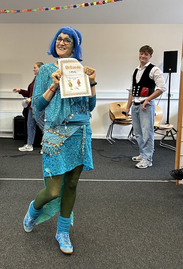 Eine Frau in einem blauen, funkelnden Kostüm mit grünen Strumpfhosen hält stolz eine Urkunde mit der Aufschrift "1. Platz" in die Kamera. Sie trägt eine blaue Perücke und ein passendes Outfit mit Glitzerverzierungen. Im Hintergrund lächelt eine Person in Trachtenkleidung.