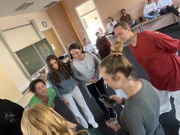 Gutgelaunter gemeinsamer Start ins Schuljahr Schülerinnen und Lehrkräfte stehen im Kreis zusammen leicht vorgebeugt als hörten sie aufmerksam zu. Eine Schülerin hält ein Handy und einen Stapel Karten in den Händen.