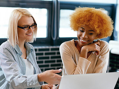 Zwei Personen sitzen an einem Tisch und arbeiten zusammen an einem Laptop. Eine Person hat kurze blonde Haare und trägt eine Brille, die andere hat voluminöse, lockige Haare und trägt einen beigen Pullover. Beide lächeln und wirken konzentriert.