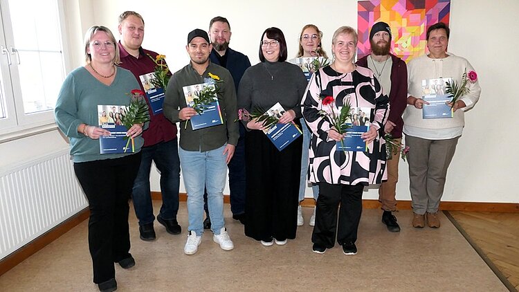 Die neun Absolventinnen und Absolventen der Klasse EZ 22b posieren, mit Abschlusszeugnis und roten und gelben Gerbererblumen in der Hand, für ein gemeinsames Erinnerungsfoto.