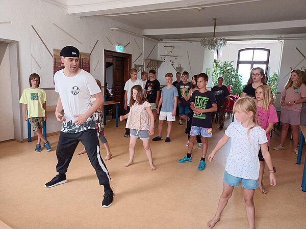 Sprachprojekte mit internationalem Flair Jungen und Mädchen stehen in einer kleinen Turnhalle in Reihen angeordnet und machen auf einem Parkettboden Tanzübungen mit ihren Beinen und Armen.