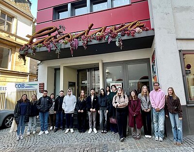Eine Gruppe von 16 jungen Erwachsenen steht lächelnd vor dem Eingang eines Gebäudes mit dem Schriftzug 'Casino' über der Tür. Das Gebäude ist mit einer dekorativen Blumen- und Pflanzenkette geschmückt.