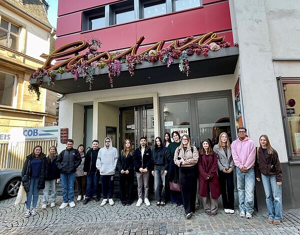 Eine Gruppe von 16 jungen Erwachsenen steht lächelnd vor dem Eingang eines Gebäudes mit dem Schriftzug 'Casino' über der Tür. Das Gebäude ist mit einer dekorativen Blumen- und Pflanzenkette geschmückt.