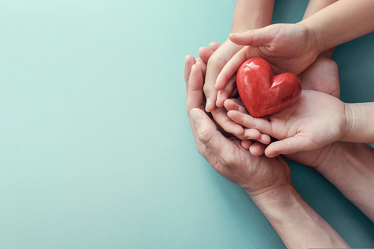Spendenlauf für Hospiz wird zum vollen Erfolg Ein rotes Herz wird von drei Handpaaren gehalten. Es könnte sich um eine Familie, die aus Vater, Mutter und Kind besteht, handeln. Das Bild symbolisiert Fürsorge.