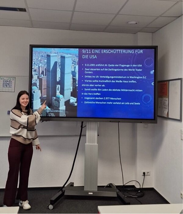 Eine Schülerin präsentiert eine Folie zum Thema „9/11 – Eine Erschütterung für die USA“. Der Bildschirm zeigt ein Bild der Zwillingstürme des World Trade Centers und einen Text mit Fakten zur Anschlagsserie.