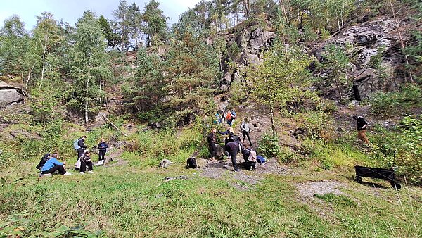 Eine Gruppe von Menschen verteilt sich auf einer grünen Wiese vor einem steilen, bewaldeten Felsen. Einige sitzen, andere steigen den Hang hinauf.