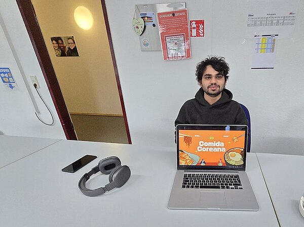 Ein Azubi sitzt an einem weißen Tisch. Vor ihm steht ein aufgeklappter Laptop, dessen Bildschirm eine orangefarbene Grafik mit dem Text 'Comida Coreana' zeigt. Auf dem Tisch liegen ein Smartphone und große, ohrumschließende Kopfhörer. Im Hintergrund hängen an der Wand verschiedene Aushänge, darunter ein Feuerlöscher-Hinweis und ein Kalender. Die Tür zum Flur steht offen, und an der Wand im Flur hängt ein gerahmtes Foto.