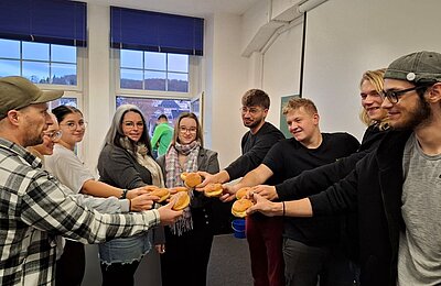 Mehrere Personen stehen im Kreis und halten jeweils einen Berliner beziehungsweise Pfannkuchen in den Händen, die sie in die Mitte strecken.