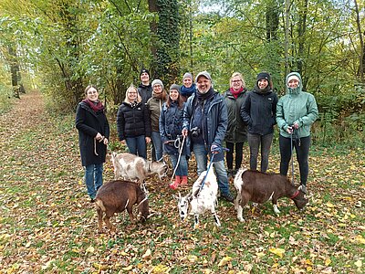 Elf Personen stehen auf einem herbstlichen Waldweg; mehrere halten Ziegen an Leinen, vier kleine Ziegen grasen im Vordergrund.