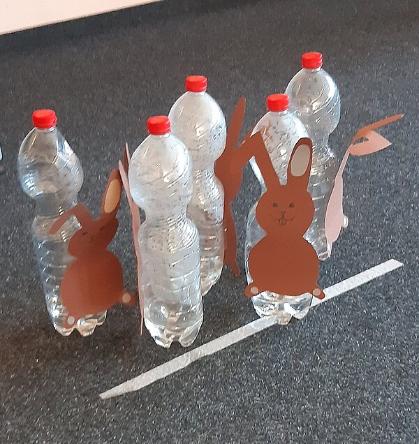 Einstimmung auf Ostern Fünf minimal mit Wasser befüllte 1 Liter Flaschen aus Plastik sind mit selbst gebastelten Hasen aus braunem Tonpapier hinter einer weißen Linie auf dem Boden in kegelähnlicher Form platziert.
