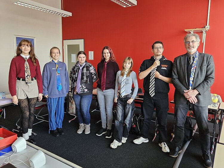 Eine Gruppe von Schülerinnen und Schülern der Euro Akademie Bamberg steht in einem Klassenraum vor einer roten Wand und präsentiert stolz ihre selbst gebundenen Krawatten. Die Auszubildenden haben im Englischunterricht das Binden von Krawatten geübt und zeigen unterschiedliche Stile und Farben. Ihr Englischlehrer steht ebenfalls mit ihnen in der Reihe und alle posieren gut gelaunt für ein Gruppenbild.