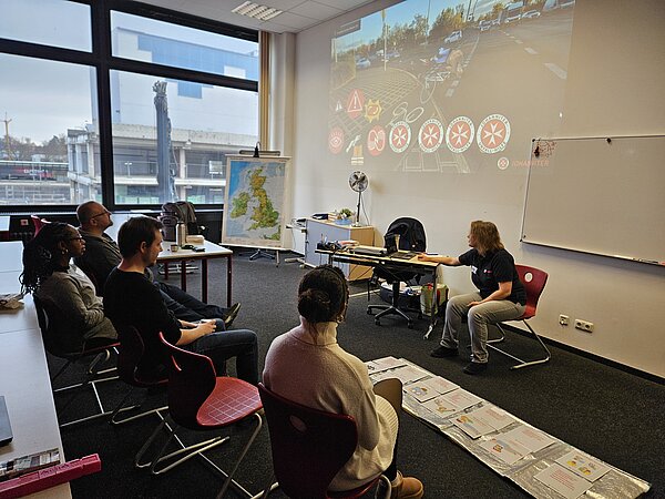Erste-Hilfe-Schulung im Seminarraum: Trainerin zeigt eine Folie mit Verkehrsunfall-Symbolen, fünf Teilnehmende sitzen zugewandt; auf dem Boden liegen Ablaufkarten, neben der Leinwand steht ein Flipchart.