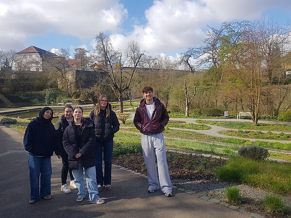 Fünf Jugendliche stehen in einer Gartenanlage auf einem Weg und posieren vor Beeten, Bäumen und einer alten Mauer.