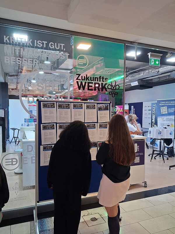 Zwei junge Frauen stehen vor einer Tafel mit Aushängen. Dahinter sind eine Glaswand und Messestände zu sehen.