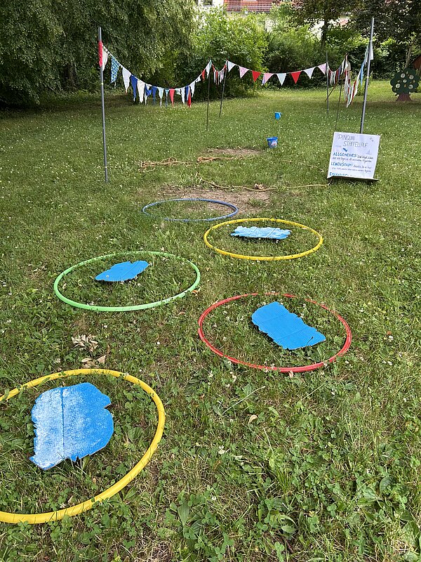 Großer Spielparcours auf einer Wiese vor dem Gebäude des Freizeitzentrums Pößneck. Der Parcours ist mit bunten Wimpelketten, Hula-Hoop-Reifen, kleinen Hindernissen und weiteren Spielmaterialien aufgebaut.