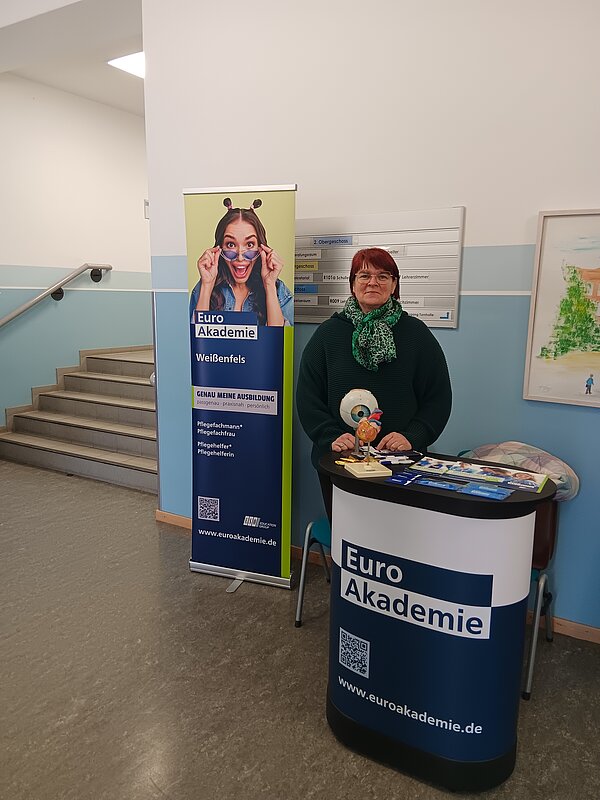 An einem Messestand der Euro Akademie steht eine dunkel gekleidete Frau mit kurzen roten Haaren, die in die Kamera lächelt.