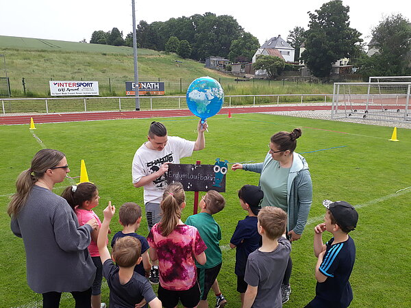 Unser Motto zum Sportfest: "Auf zu neuen Galaxien!" Ein Mann hält einen blauen Globus-Ballon, während er einer Gruppe Kinder und zwei Erwachsenen etwas erklärt. Das Schild trägt die Aufschrift „Erdumlaufbahn“.
