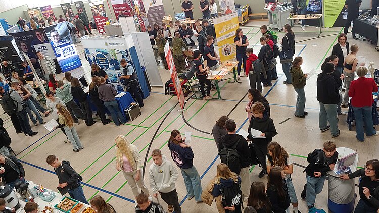 In einer Sporthalle tummeln sich zahlreiche Schüler um verschiedene Berufsinfostände.