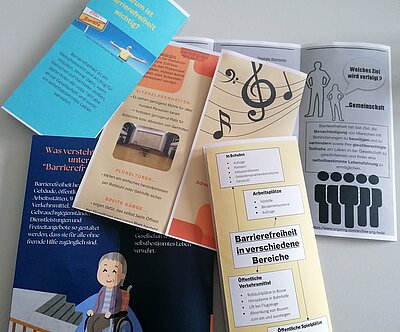 Flyer mit orangenen oder blauen Hintergrund liegen auf einer weißen Tischplatte. Inhaltlich wurden die Flyer von den Sozialassistent*innen erarbeitet und handeln von Barrierefreiheit.