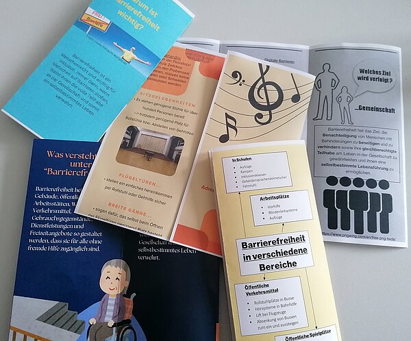 Praktikumsvorbereitungen der Sozialassistent*innen Flyer mit orangenen oder blauen Hintergrund liegen auf einer weißen Tischplatte. Inhaltlich wurden die Flyer von den Sozialassistent*innen erarbeitet und handeln von Barrierefreiheit.