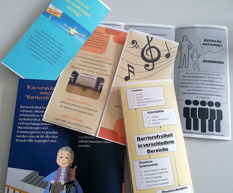 Praktikumsvorbereitungen der Sozialassistent*innen Flyer mit orangenen oder blauen Hintergrund liegen auf einer weißen Tischplatte. Inhaltlich wurden die Flyer von den Sozialassistent*innen erarbeitet und handeln von Barrierefreiheit.