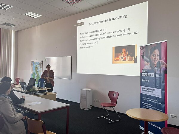 Eine Referentin stellt in einem Seminarraum Studien- und Berufsperspektiven im Bereich Übersetzen und Dolmetschen vor; daneben steht ein Roll-up der Euro Akademie Bamberg.