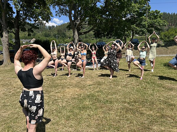 Auf einer Wiese praktiziert eine Gruppe junger Menschen Yoga: sie alle stehen auf einem Bein, halten das andere Bein angewinkelt und halten die Arme im Bogen über ihre Köpfe.