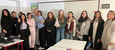 Eine Gruppe junger Frauen posiert lächelnd für das Gruppenfoto im Klassenraum vor einer großen weiße Magnettafel. Eine Person hält einen kleinen Weihnachtsbaum aus Pappe in den Händen.