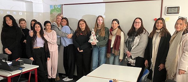 Eine Gruppe junger Frauen posiert lächelnd für das Gruppenfoto im Klassenraum vor einer großen weiße Magnettafel. Eine Person hält einen kleinen Weihnachtsbaum aus Pappe in den Händen.