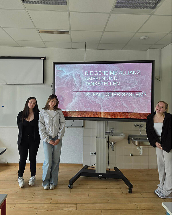 Drei Personen stehen vor einem Bildschirm, auf dem der Titel „Die geheime Allianz: Ampeln und Tankstellen – Zufall oder System?“ von Amely, Anna und Laura zu sehen ist.